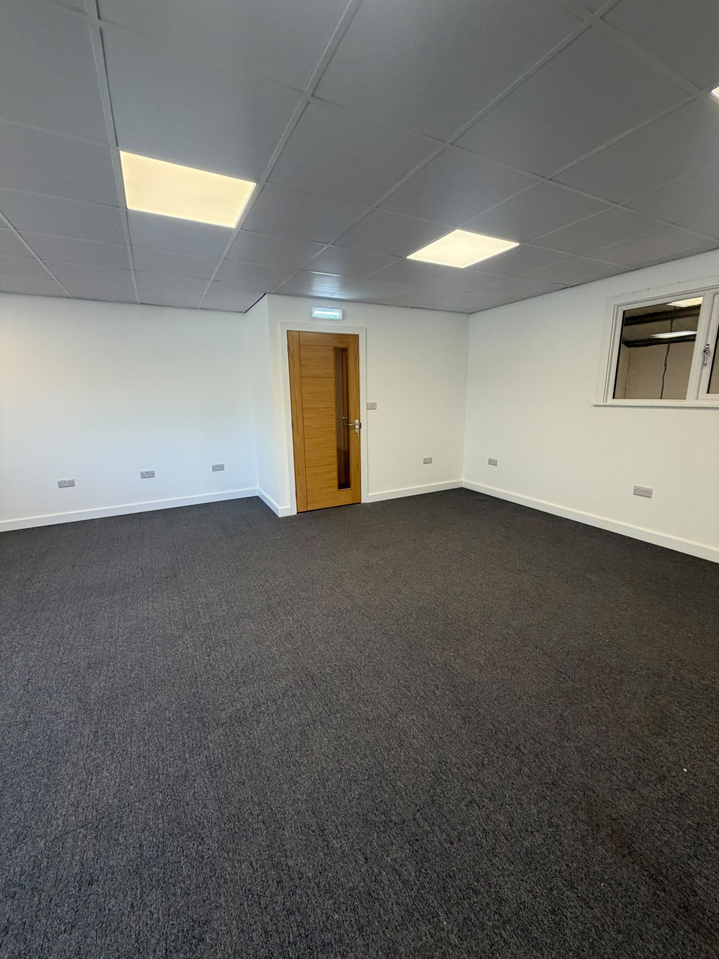 Empty first-floor office with grey carpet tiles, LED ceiling panels and oak door